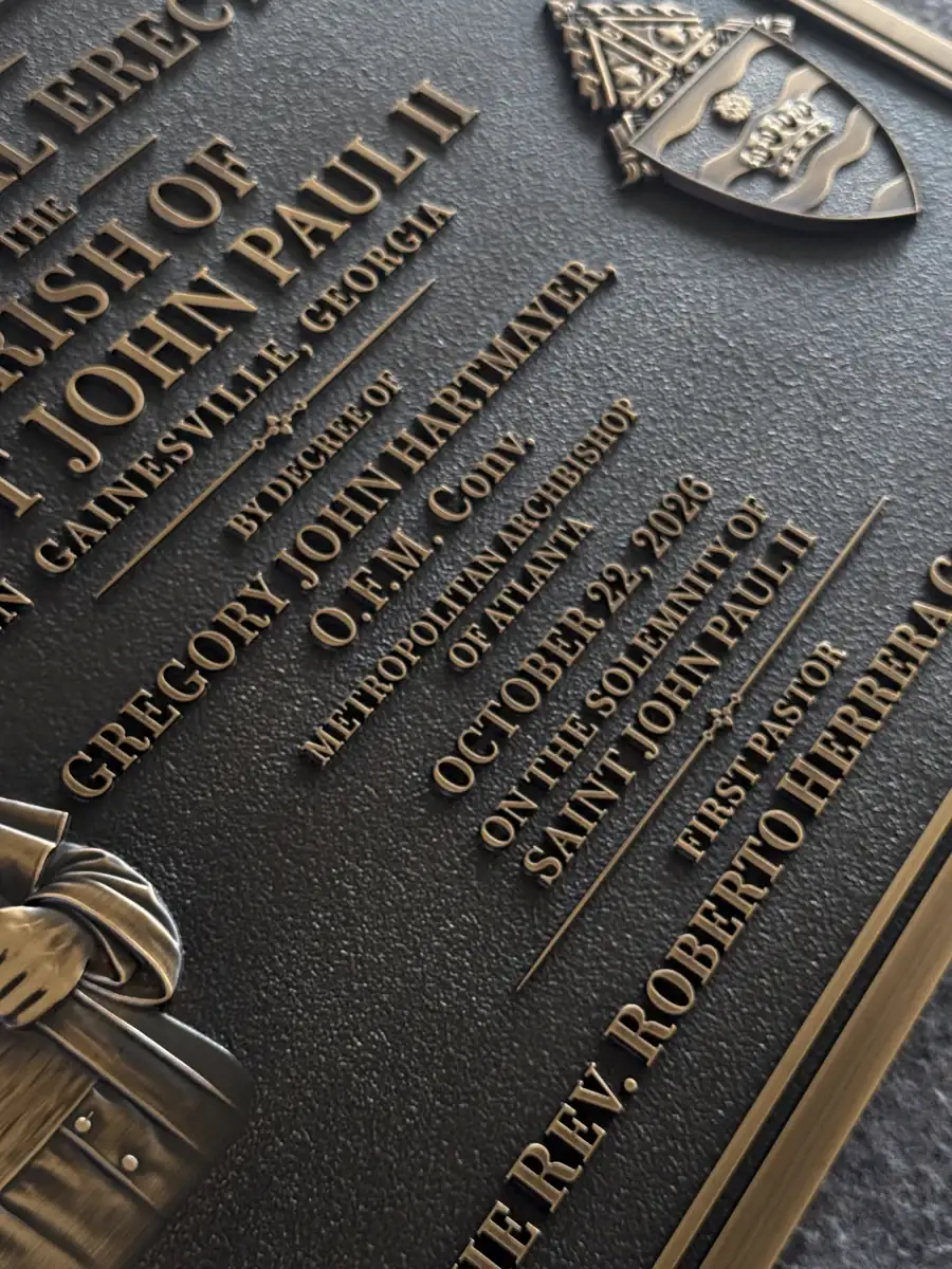 Close-up of raised lettering, dark background texture and a coat of arms on the solid copper church plaque